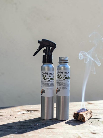 Two silver metal bottles of Black Apothecary Palo Santo incense sticks, one with a black spray trigger top and one with a silver cap, displayed on a rustic wooden surface with a smoldering incense stick and smoke rising against a neutral background.